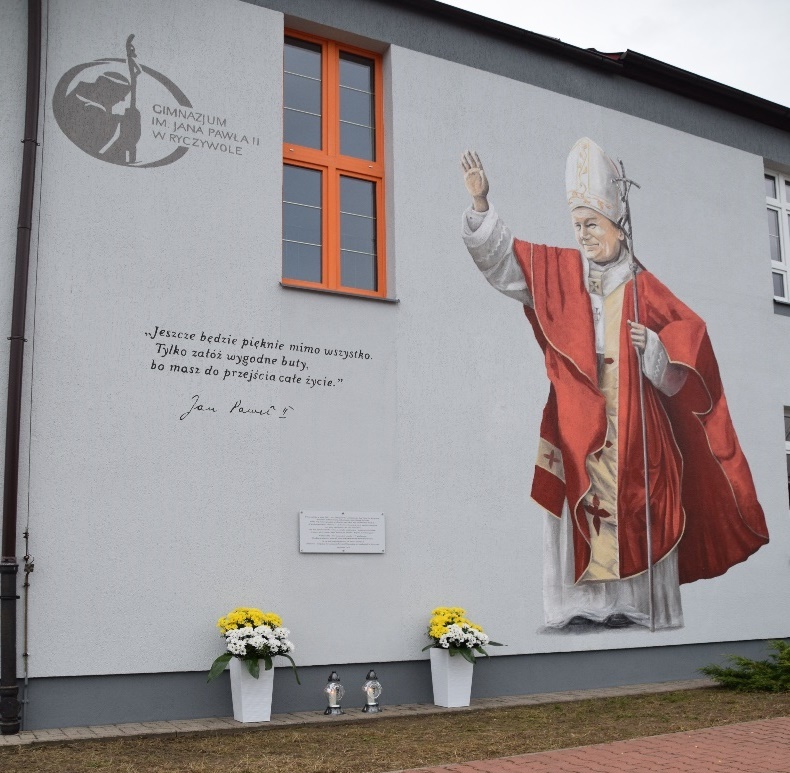 Zdjęcie przedstawia widok na ścianę Szkoły Podstawej w Ryczywoile, po prawej stronie widać mural, na którym znajduje się Jan Paweł II z uniesioną ręką po lewej stronie znajduje się tablica pamiątkowa oraz cytat i logo Gimnazjum im. Jana Pawła II w Ryczywole